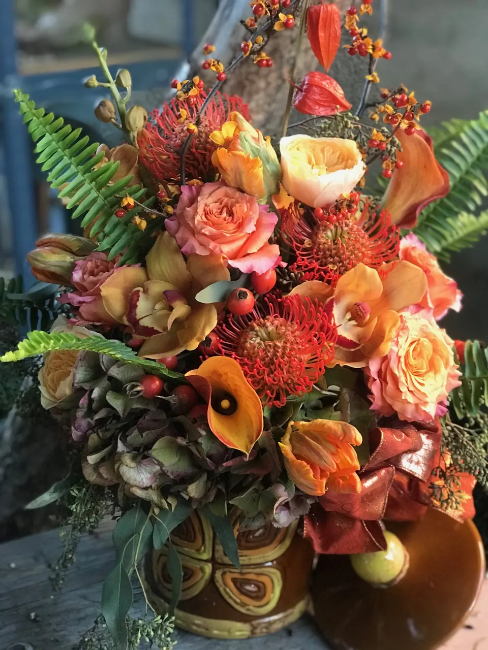 Autumn floral arrangement with orange roses, burgundy accents, pods, and sculptural foliage.