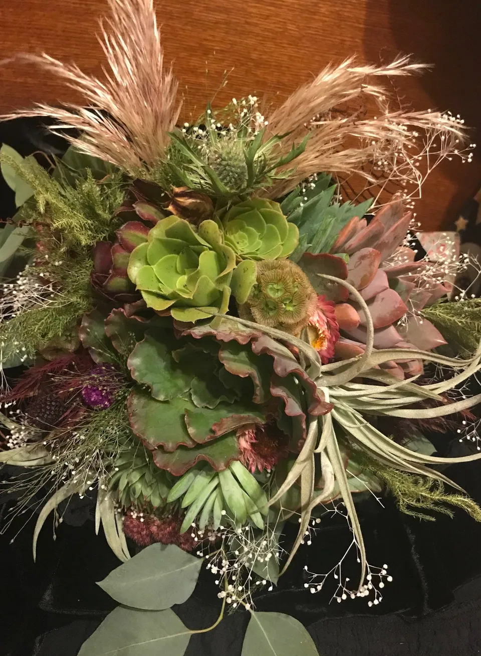 Unusual floral piece with succulents, pale roses, feathery texture, and deep purple accents.